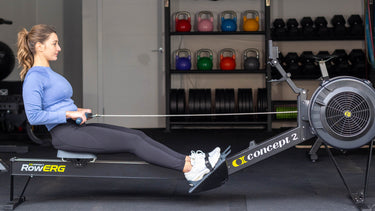 Woman using a rowing machine in a home gym with various fitness equipment in the background.