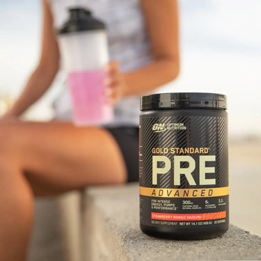 Woman sitting on low wall at beach with shaker in hand and tub of pre-workout beside her.