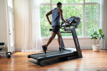 Person using a treadmill in a home setting with large windows and a plant.